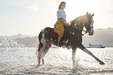 Caucasian blonde girl riding a horse through the water at sunrise
