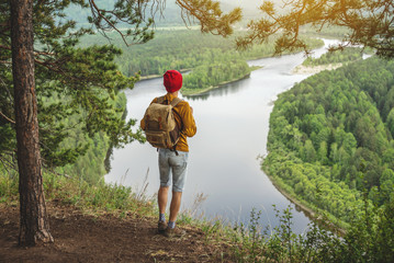 Traveler with a backpack is standing on edge of a cliff and is looking on a green valley. Freedom and unity with nature