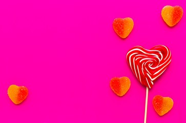 Fototapeta premium Striped screwed red and white heart-shaped candy on a stick and marmalade in sugar on a pink background. Sweet treat. A delicious gift. Use for postcard. Love concept. Valentine's Day.