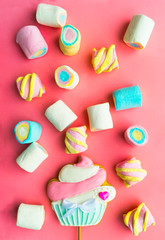 Scattered multi-colored marshmallows and gingerbread on a stick in the form of a muffin on a pink background. Holiday treat. Children's holiday. Use for postcards. Vertical layout. Selective focus.