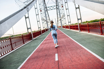Back view of running girl. Young athlete woman is jogging on the pedestrian bridge. Outdoors run