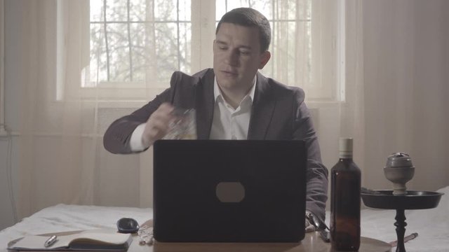 Young relaxed man drinking whiskey after online chat and leaning back on bed. Portrait of lazy Caucasian freelancer on remote work during Covid-19 lockdown. Lifestyle, freelance. S-log-2 ProRes 422 HQ