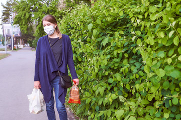 A young girl in a blue jacket stands next to green shrubs in a medical mask and looks to the left