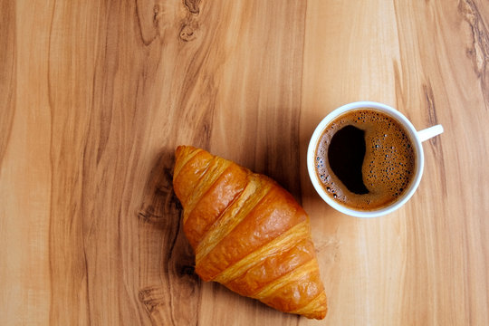 Top view shot of appealing plain mini croissant baked to golden crisp and cup of black coffee on exotic wood textured backgroud. Traditional french pastry. Close up, copy space.