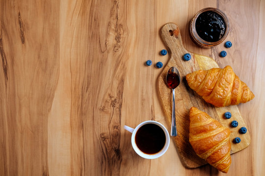 Top view shot of appealing plain mini croissant baked to golden crisp, cup of black coffee & blueberry jam on exotic wood textured backgroud. Traditional french pastry. Close up, copy space.