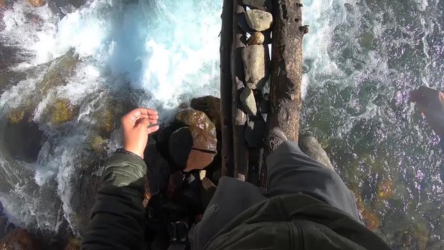 An amateur, solo backpacker attempting to cross a dangerous, fast flowing mountain river stream by slowly stepping on a wooden log. Shot from hiker&rsquo;s point of view.