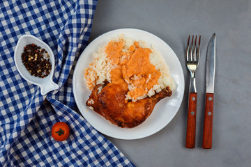 Close up view of chicken curry leg with rice served on the white plate on the grey background. Blue napkin, fork, knife, pepper decorate composition. Nutritious food, appetizing dish for lunch. 
