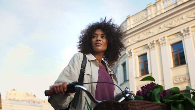 Afro american model with curly hair, in casual outfit. Smiling, looking around, holding her bicycle by handlebar, walking surrounded by buildings