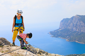 Young mother, child in safety equipment climb to mount top by via ferrata beginner route. Family...