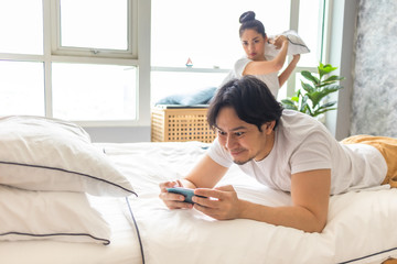 Excited Asian man plays mobile game and angry woman standing behind him.