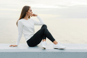 Young woman in white sweater and black leggings is resting after jog on promenade