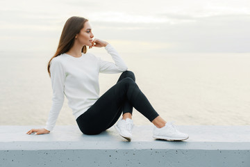 Young woman in white sweater and black leggings is resting after jog on promenade © Mirrorstudio