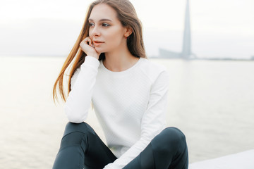 Young woman in white sweater and black leggings is resting after jog on promenade