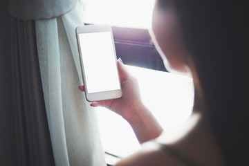 Closed up image of a woman using a white blank screen smartphone. Background of a window.