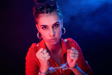 a bright brunette girl in a red jumpsuit handcuffed on a dark background in neon light
