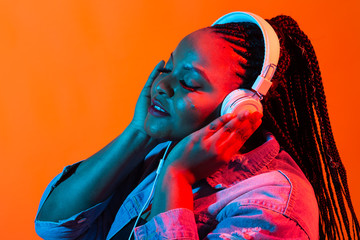 African american young woman listening to music online dancing and singing with headphones, neon...