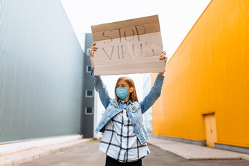 woman in a medical protective mask holds a cardboard placard with the words STOP the virus, on a street in the city