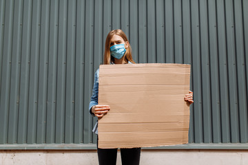 woman in a medical protective mask holds an empty cardboard poster for a copy of the space, standing in the city against a gray wall. Quarantine, coronavirus