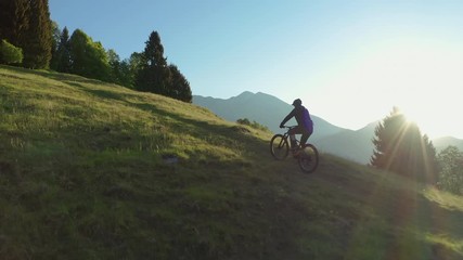 Drone is flying along an athletic man pedalling an MTB E-bike up a steep grassy hill. Beautiful view of the mountains at sunrise/sunset with sun flare. Alone in nature, thinking about life. - Powered by Adobe