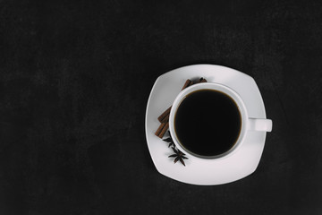 White cup with coffee and saucer, with anise star and cinnamon sticks, on dark textured background. A good idea for the menu or signboard of a restaurant or cafe, cafeteria. Top view, space for text