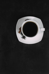 White cup with coffee and saucer, with anise star and cinnamon sticks, on dark textured background. A good idea for the menu or signboard of a restaurant or cafe, cafeteria. Top view, space for text