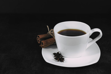White cup with coffee and saucer, with anise star and cinnamon bunch, on dark textured background. A good idea for the menu or signboard of a restaurant or cafe, cafeteria. Side view, space for text
