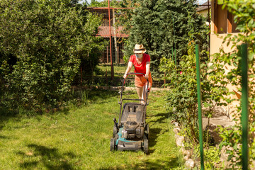 Home life during quarantine or self-isolation. A woman in her backyard mowing grass with a lawn mower on a sunny day at home wearing a surgical mask because of the coronavirus epidemic.