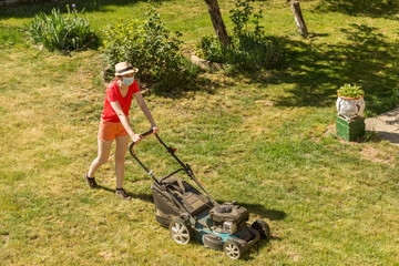 Fototapeta na wymiar Home life during quarantine or self-isolation. A woman in her backyard mowing grass with a lawn mower on a sunny day at home wearing a surgical mask because of the coronavirus epidemic.