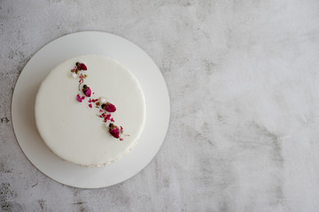 Top view of white mousse cake with smooth glaze decorated with dried rose buds  on gray textured background. Сoncise simple design, Copy space