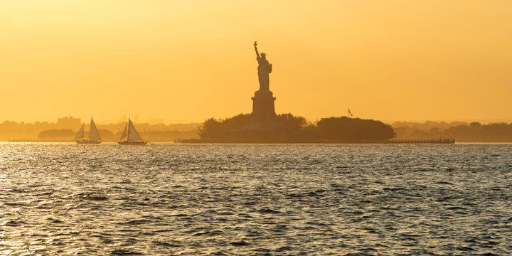 Statue of Liberty and Liberty Island across New York Harbor with sailboats in Summer sunset light. New York City (UNESCO World Heritage)