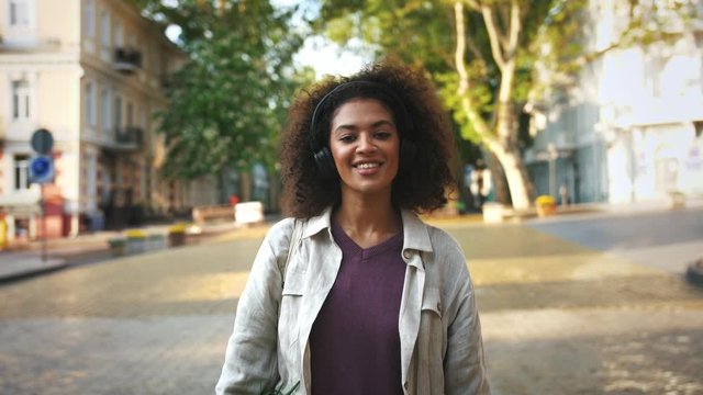 Afro-american lady in headphones, casual clothes. She smiling, dancing, listening to the music while walking by deserted street of a city. Close up