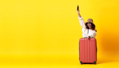 Cheerful young african woman dressed in summer clothes holding passport with suitcase over yellow banner background with copy space.