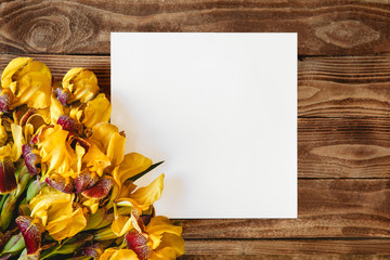 A bouquet of blooming yellow irises and white sheet of paper on a wooden background. Flat layout with space for text