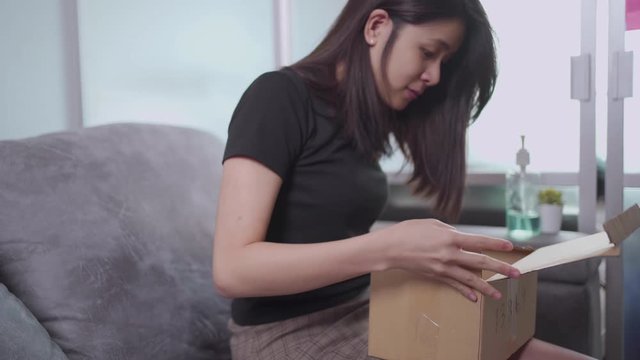 Young asian female opening small cardboard box pakage delivery, on sofa at home, receiver unpack small carton parcel container, covid-19 pandemic, goods supply delivery online shopping,  e-commerce