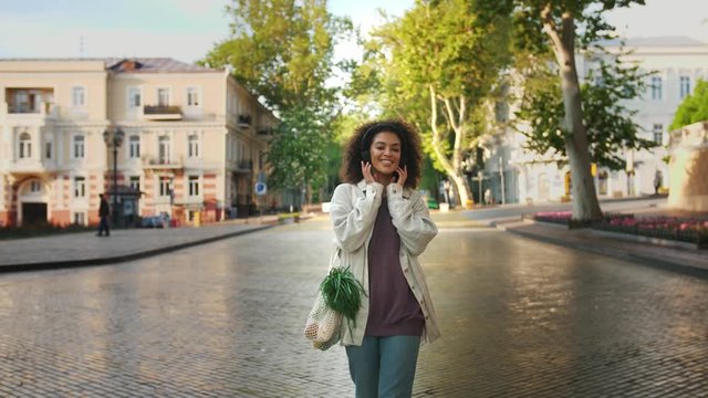 woman in headphones and casual clothes. She smiling, listening to the music, walking by deserted avenue of city. Carrying food in eco bag