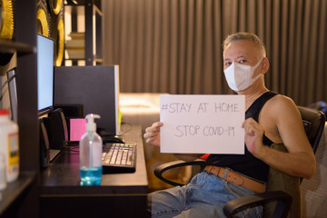 Mature Japanese man with mask showing Stay At Home sign while working overtime at home