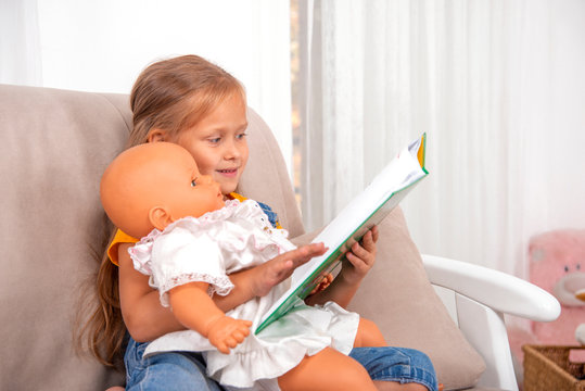 A little girl sitting on the sofa   and reads book with her doll