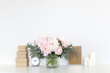 Peonies in the interior on the table, clock alarm clock, craft boxes, candles