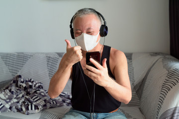 Happy mature Japanese man with mask video calling with headphones and giving thumbs up at home