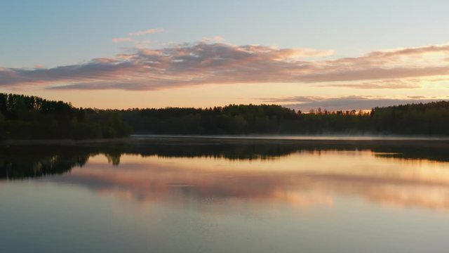 Flying over lake at sunrise. Clouds of fog above water surface. Early morning mist haze. Haze over river countryside. Pine trees on horizon reflected in water as in mirror. Beautiful dawn landscape