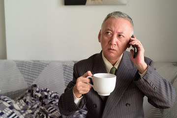 Mature Japanese businessman talking on the phone and holding coffee at home