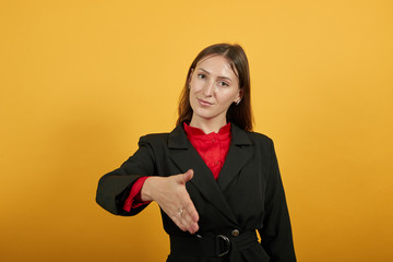 Young Attractive Brunette Woman In Black Stylish Suit, Red Shirt On Yellow Background, Friendly Female Reaches Out To Say Hello. The Concept Of Good People Etiquette, Hospitality