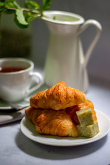 Breakfast table set up with the hot tea and croissant, cheese cubes fresh mint served in white dishes on the gray background.