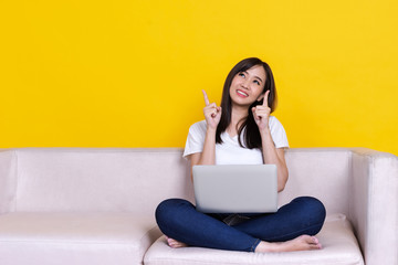 Asian woman smiling and point finger to copy space and working from home use laptop computer while sitting on sofa over isolate yellow background