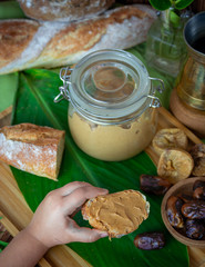 Child’s hand taking a peanut butter sandwich from the table with homemade peanut butter glass jar, fresh baguette, Turkish coffee pot, dates and figs. Close up top view shot of the breakfast table.