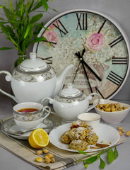 Table set up with the home made coconut cookies, white and gray print tea set, black tea, lemon, pistachios nuts, vase with the green branch, wall clocks at the back on the gray background.