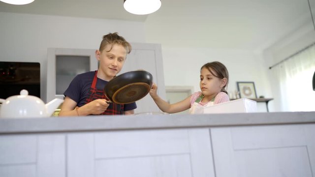 Kids Home cooking concept image. Sister and Brother dressed aprons making a homemade pancakes on the home kitchen. Girl poring a liquid dough on the hot pan. Kids on kitchen 4K concept footage.