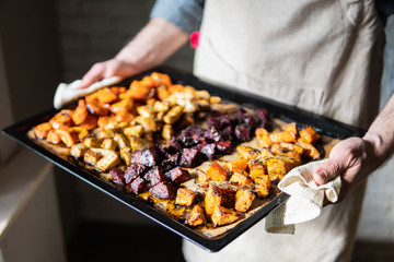 Male hands holding oven tray with vegetables mix. Vegetarian man cooking sweet potatoes and other veggies at home