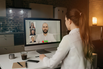 Back view of woman working remotely talking to her colleagues about business in video conference on desktop computer at home. Multiethnic business team using computers for online meeting in video call