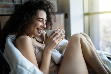 Beautiful young woman in underwear sitting next to a window and enjoying a cup of coffee in the morning